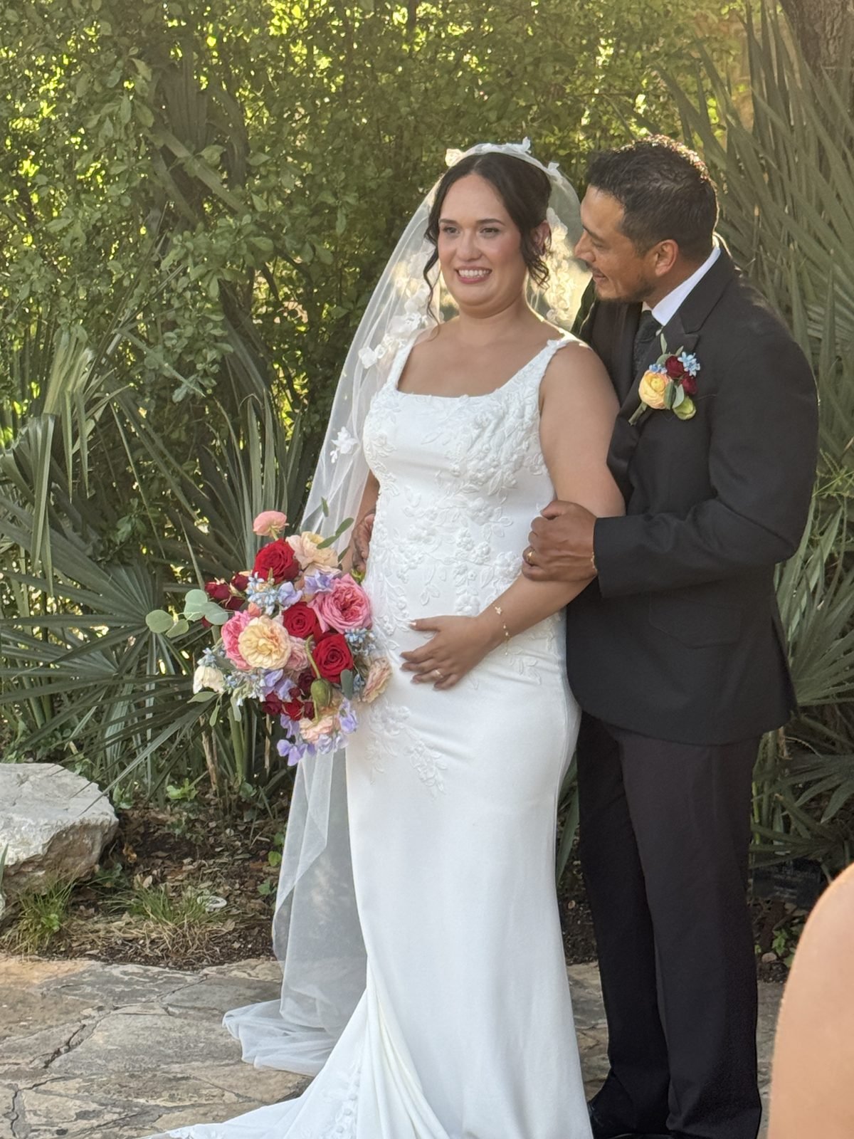 Groom in a black suit with his arm around the bride, who wears a fitted white gown and long veil and holds a colorful rose bouquet, standing among green foliage