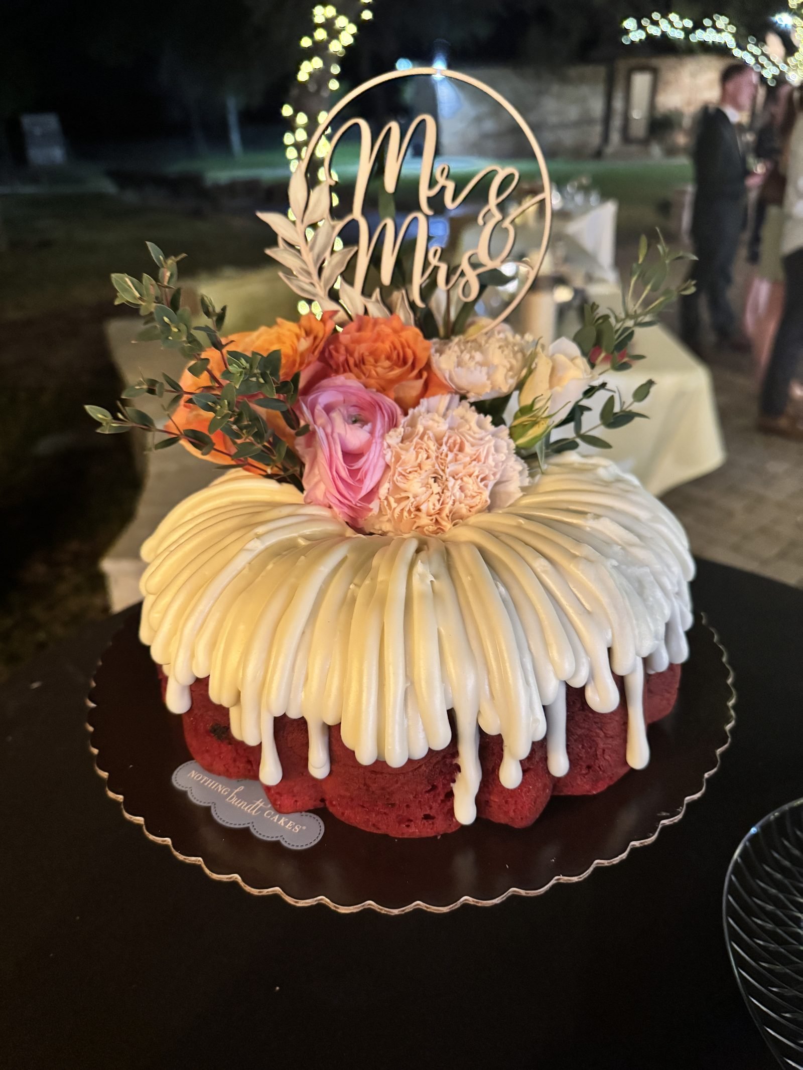 Red bundt wedding cake with white icing drizzle topped with orange and pink flowers and a gold script cake topper