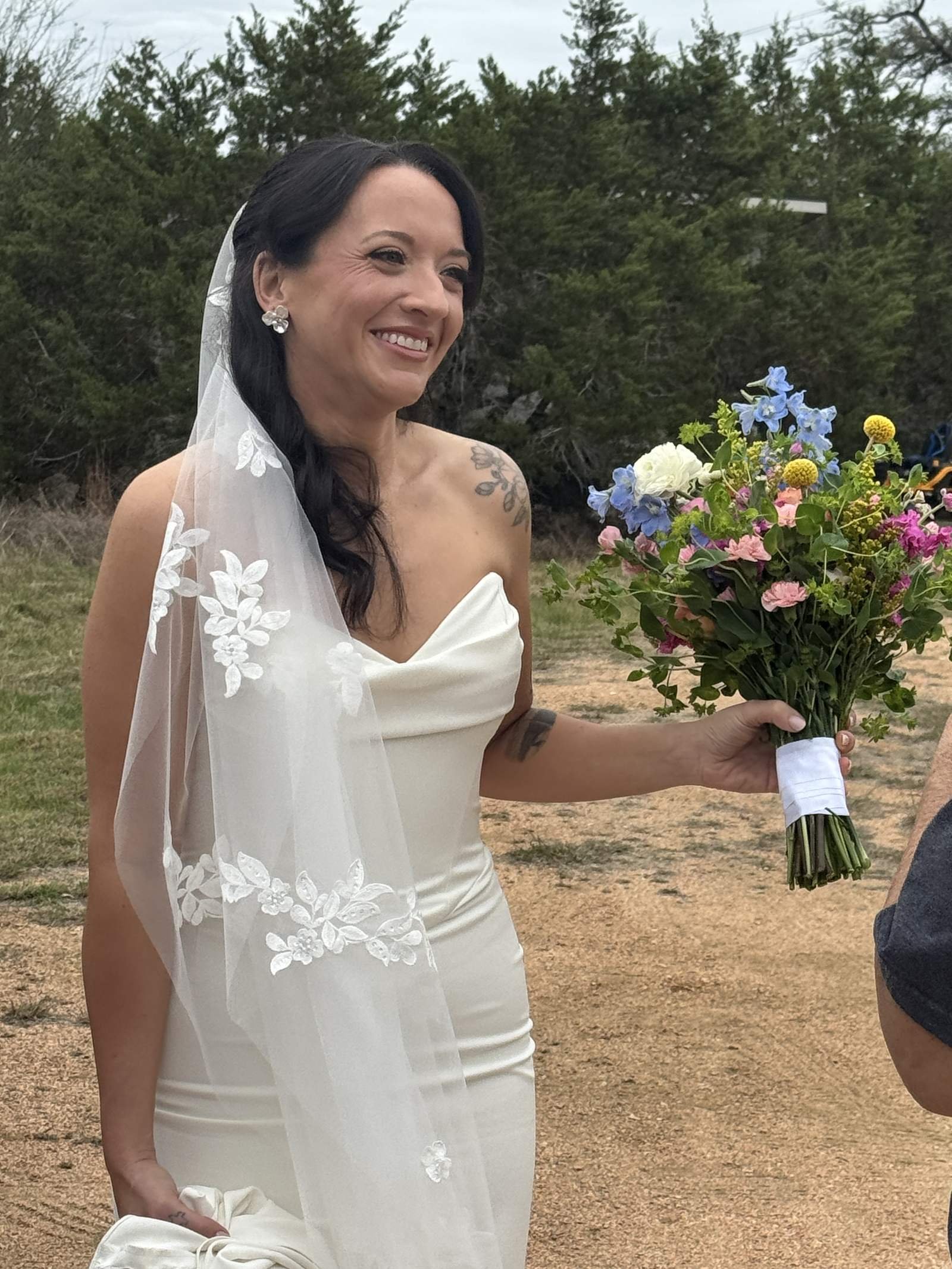 Smiling bride in a strapless gown and long veil holding out a colorful bouquet outdoors