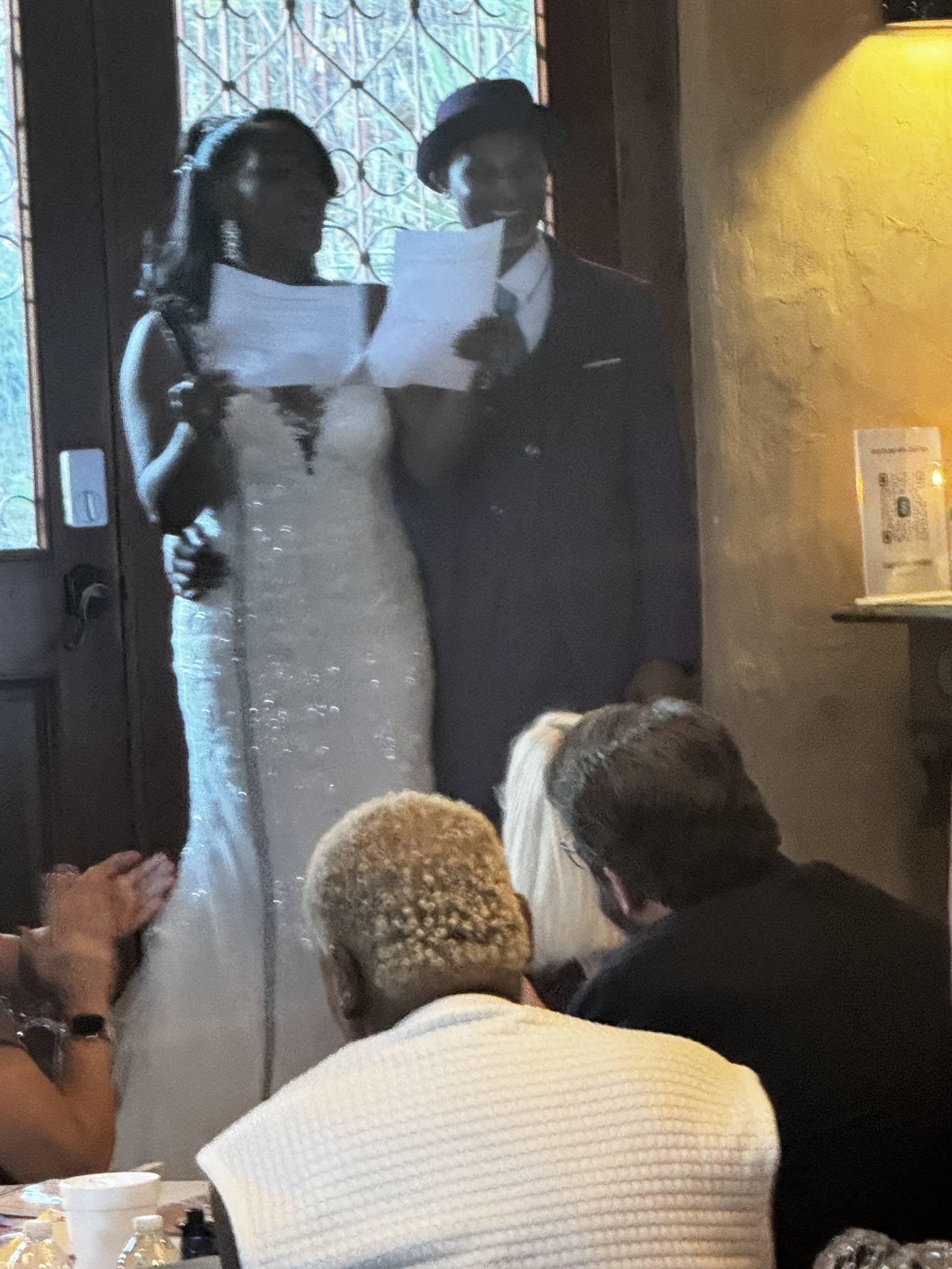 Bride in a sparkly gown and groom in a hat reading vows from paper as seated guests look on indoors