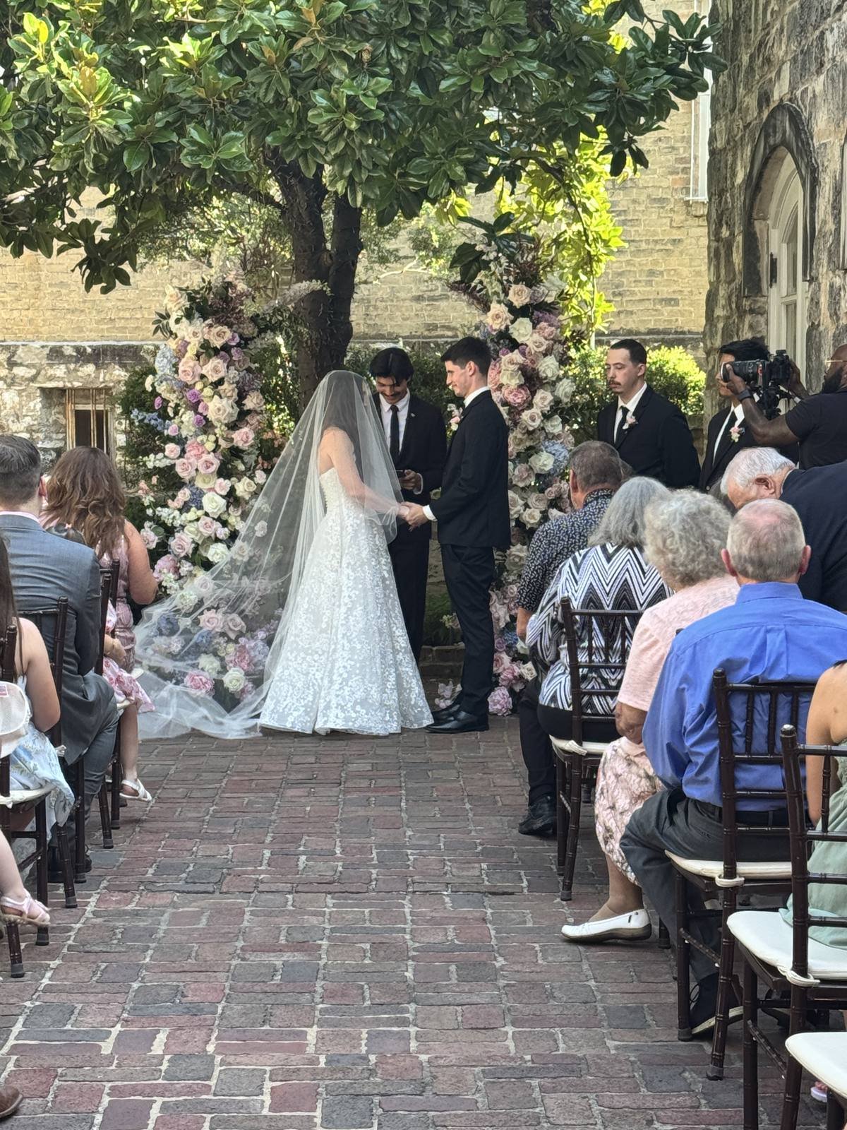Bride in a long cathedral veil holding hands with the groom at an outdoor courtyard ceremony with tall pastel floral arches as family watches from wooden chairs