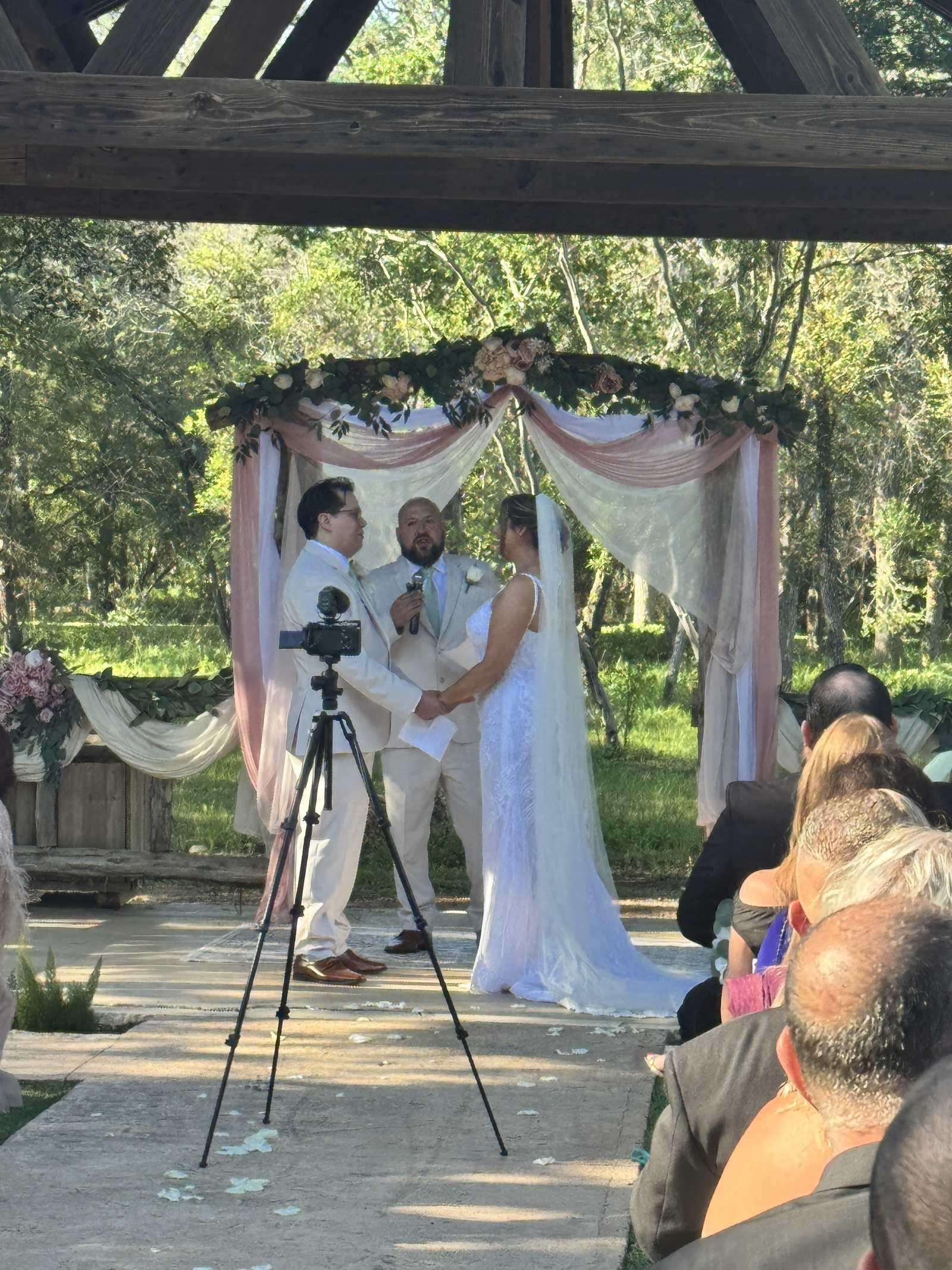 Outdoor wedding ceremony under a draped arbor with greenery, filmed by a video camera on a tripod in the aisle