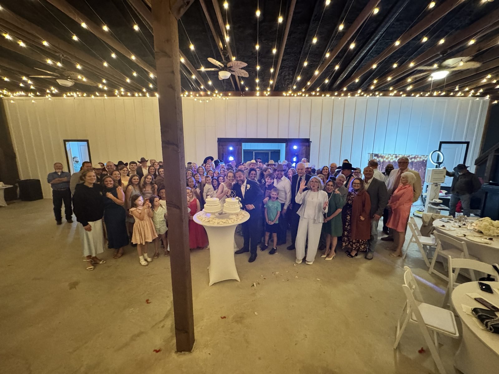 Large group of wedding guests posing with the couple in a white reception hall under rows of string lights
