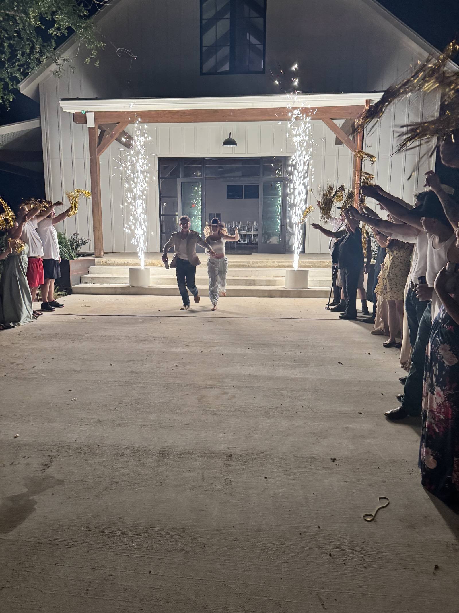 Newlyweds walking toward guests between two sparkler fountains in front of a white building at night