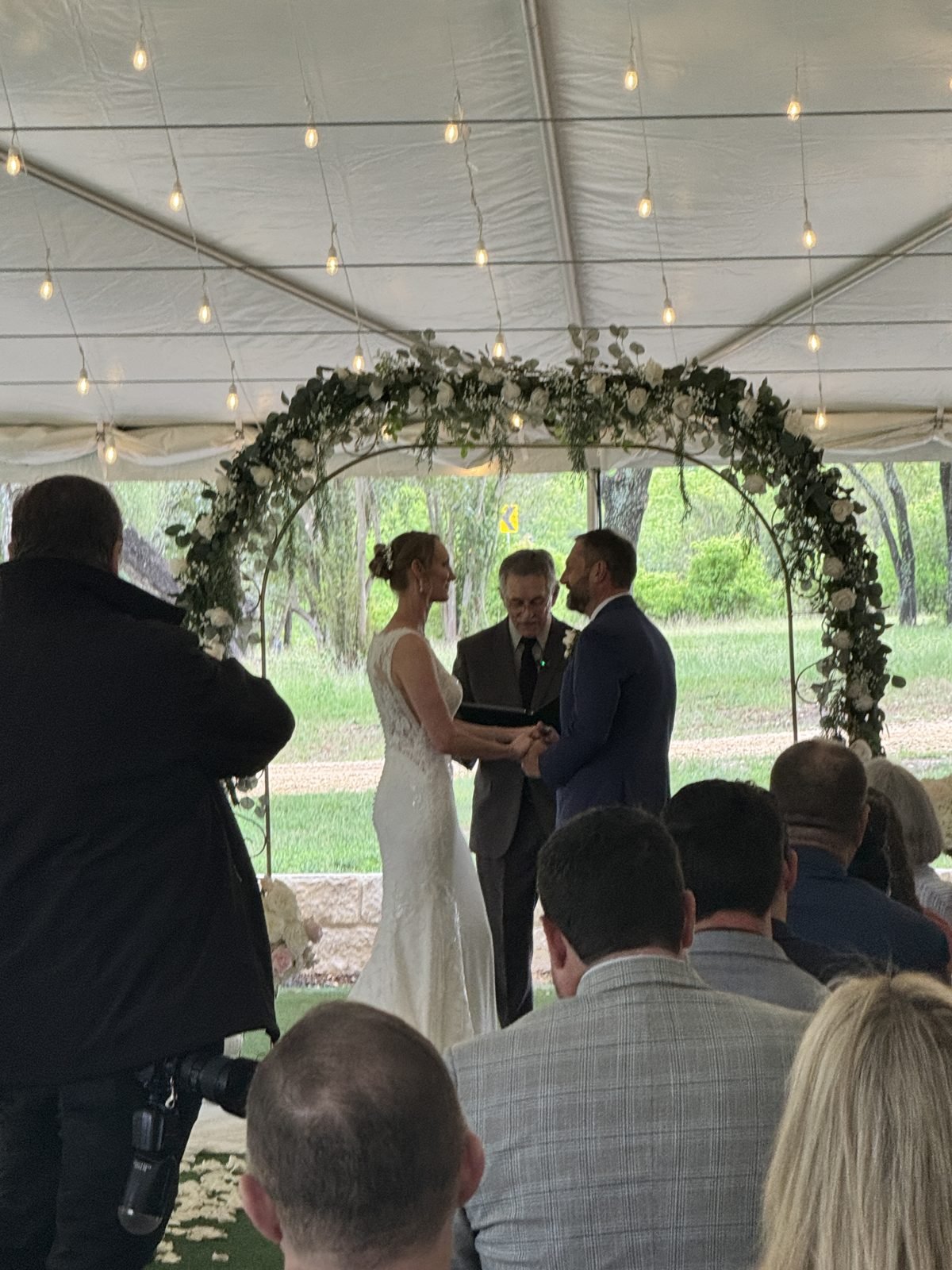 Bride and groom holding hands under a greenery and white flower arch inside a tent with string lights while the officiant reads and guests look on